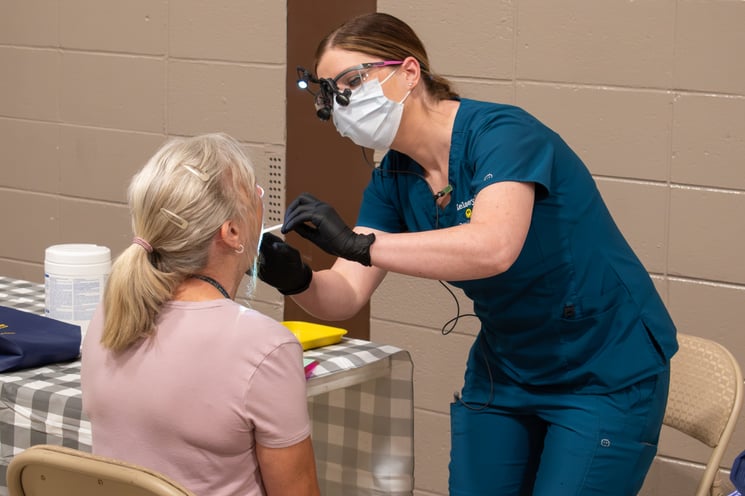 A female hygienist wearing teal scrubs, loupes and a mask bends over an older woman with ash-blond hair whose back is to the camera. The hygienist is about to conduct a screening.