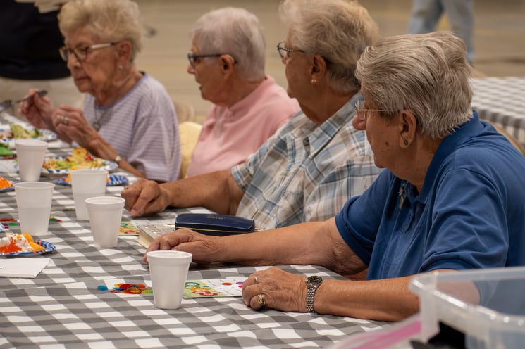 A group of older adults sitting at a table with a checked tablecloth eat lunch and play oral health bingo.