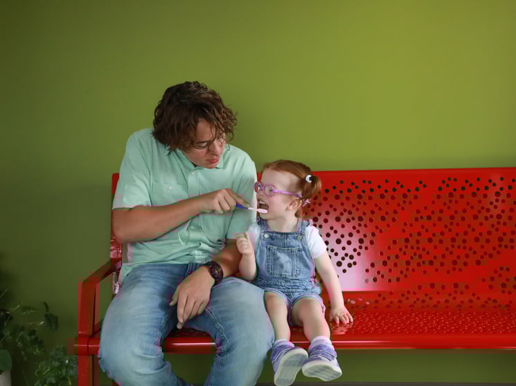 A little girl with overalls, pigtails and purple glasses opens her mouth for a toothbrush, which is held by her dad. They sit together on a red metal bench.