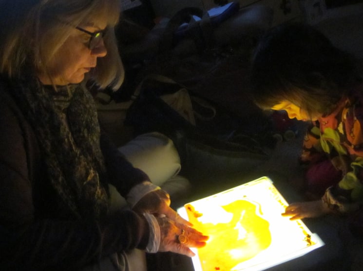 A dark photo of an older woman wearing glasses interacting with a toddler over a brightly lit lightbox.