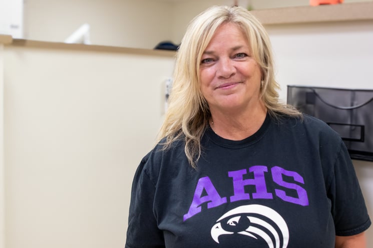 A woman with shoulder-length blonde hair wearing a black Aiken High School Falcons T-shirt smiles at the camera.