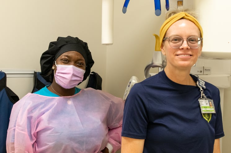 Two female dental assistants, one gowned in PPE and the other in a blue T-shirt with a yellow headband smile at the camera. They're standing in front of a panoramic X-ray machine.
