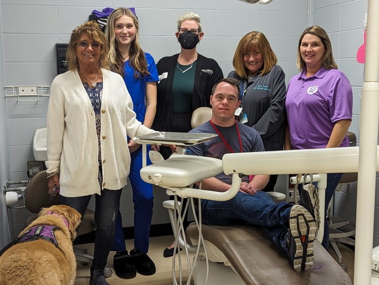 Group photo of five women standing around a man sitting in a dental chair.