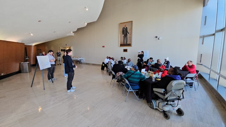 A group of people from varied racial backgrounds and abilities sits at a table while another group of people presents in front of an easel.