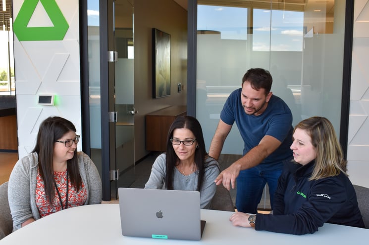 A team of three women and one man huddles around a laptop computer.