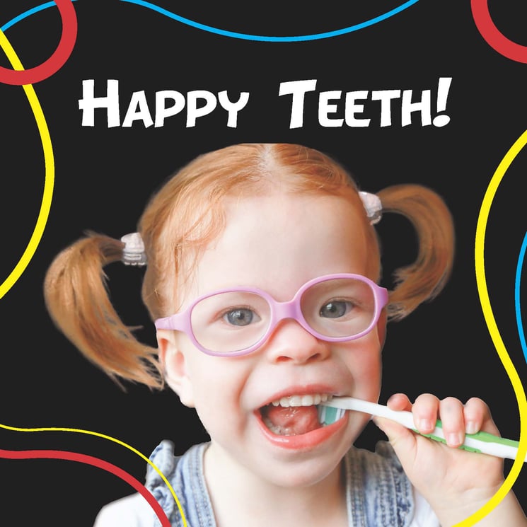 A smiling little girl with red hair in pigtails wearing pink glasses holds a toothbrush in her mouth.