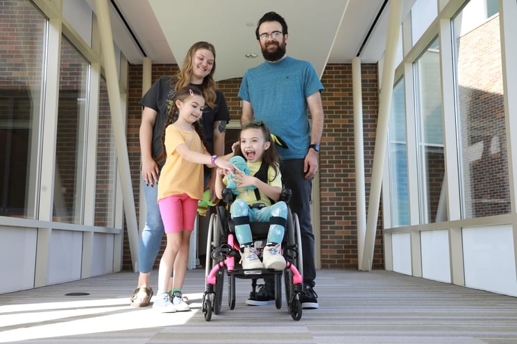 A mom and dad stand in a hallway with two young girls, one of whom uses a pink wheelchair and clutches a Squirtle plushie. Everyone is smiling.