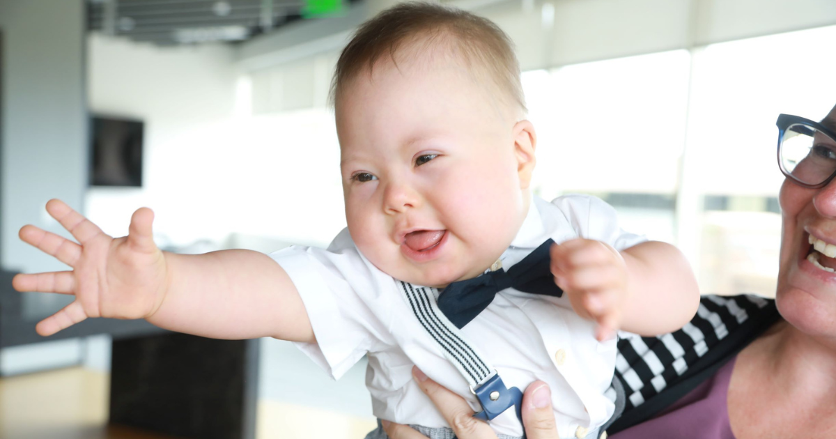 Levi, a toddler with Down syndrome, smiles and stretches his hand out as though he's reaching beyond the frame. His mom holds him, laughing.