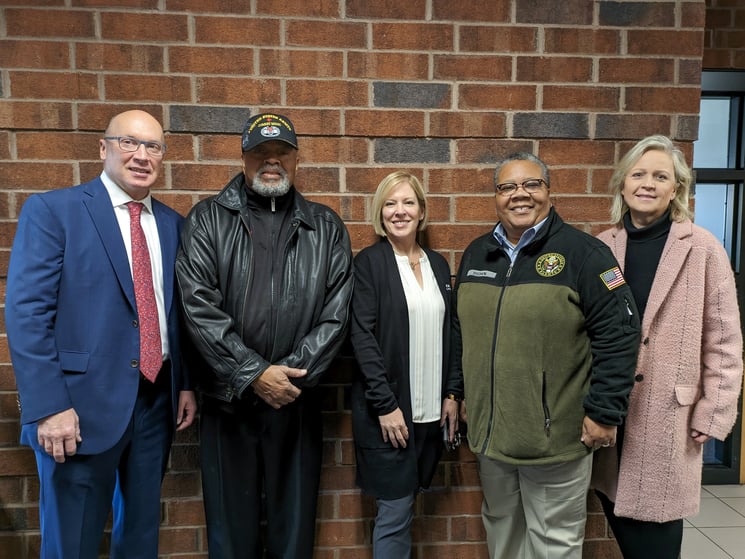 A group of five people (two men and three women) stand smiling against a brick wall.