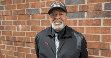 Michael Johnson, an older Black man with a salt-and-pepper beard, smiles at the camera. He's wearing a U.S. Army Combat Medic baseball cap and a black jacket.