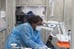 A woman with dark brown skin wearing PPE leans over a patient seated in a dental chair. The patient is just out of view, but you can see their sandals.