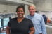 A woman with dark brown skin and braces in black scrubs smiles widely at the camera. Next to her, an older white man wearing a blue-striped collared shirt smiles with her. They're inside of a dental bus.