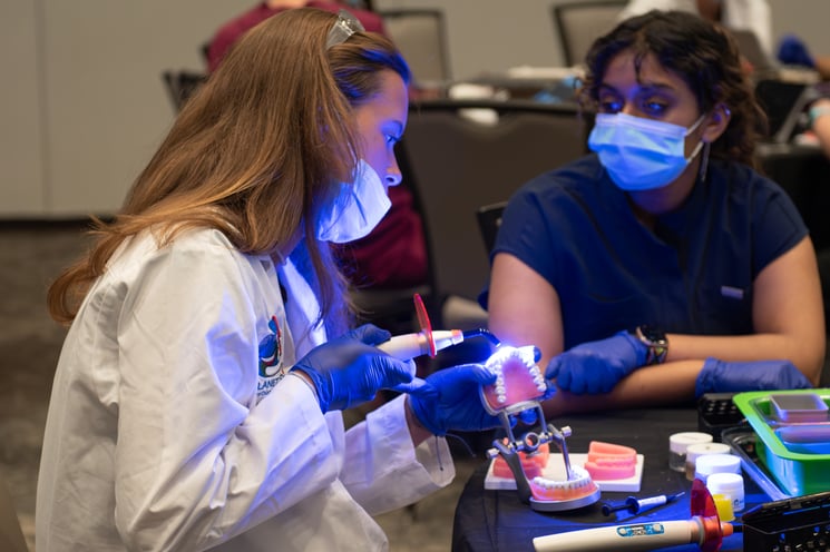 A young woman wearing a white lab coat and PPE shines a blue curing light on a mouth model while a dental student looks on
