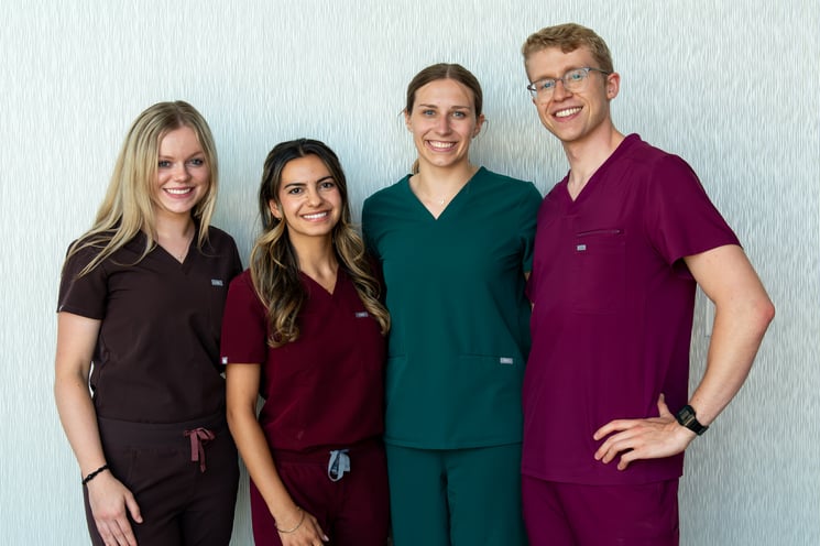 Three young women and one young man smiling and wearing scrubs of varying colors