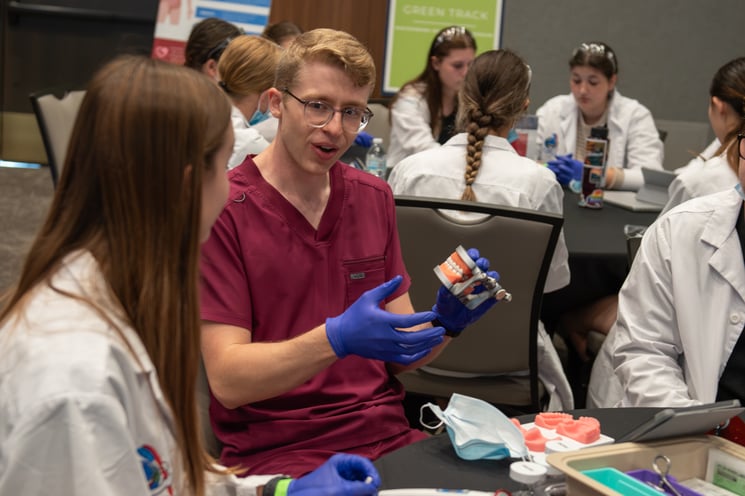A male dental student wearing maroon scrubs holds up a mouth model as he talks to high school students.