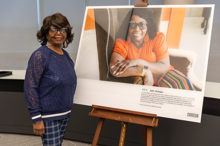 A smiling Black woman wearing fun cat-eye glasses and a blue lace blouse stands next to a picture of herself.
