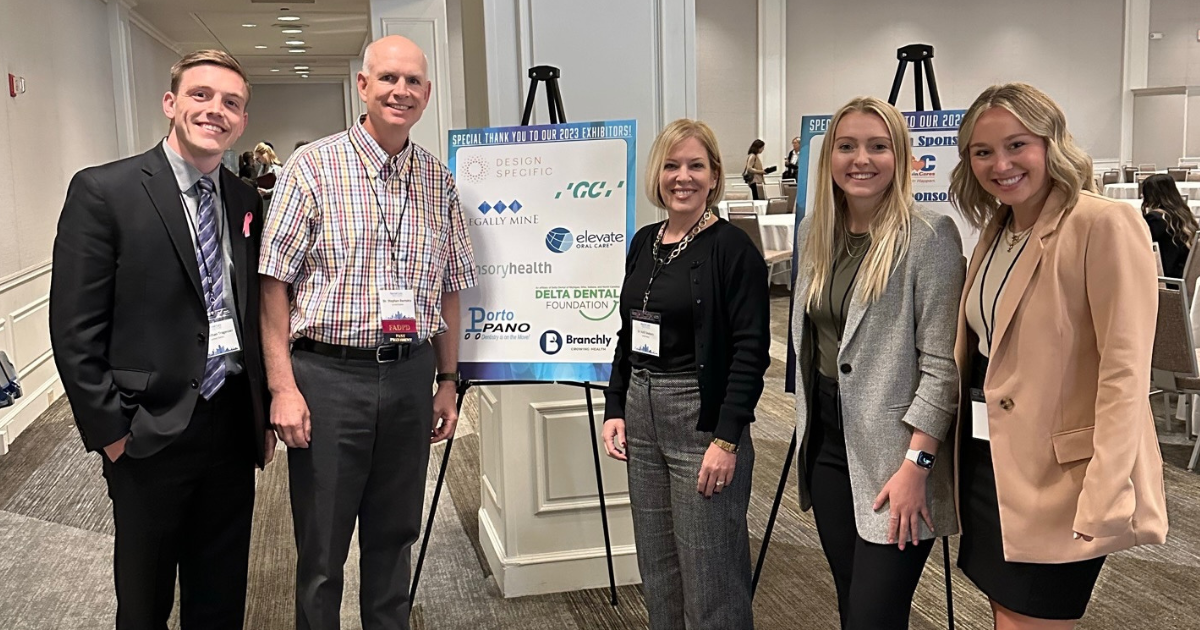 A group photo of two men and three woman standing next to a sponsor poster at a conference.