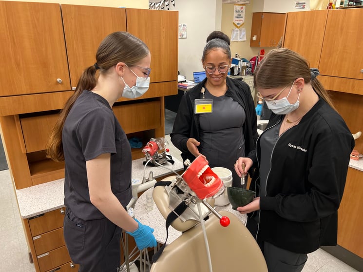 Two young women with their hair in ponytails stand over a dental mannequin, ready to do work. An older Black woman, their teacher, stands in the background observing.