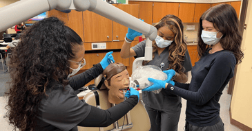 Two Black women and one White woman wearing charcoal scrubs stand around a dental mannequin holding an X-ray machine.