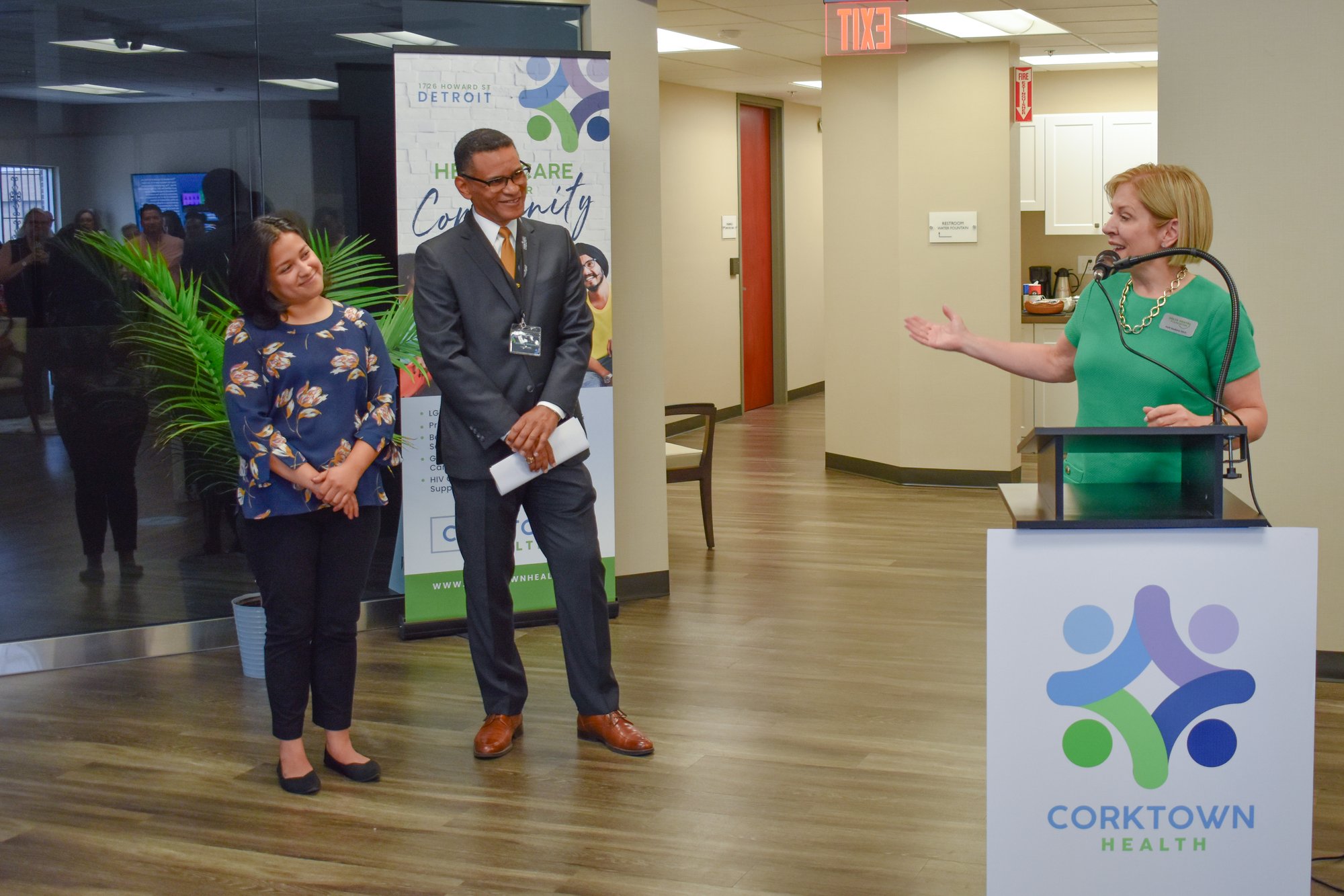 Holli Seabury stands behind a lectern with Corktown Health's logo. She gestures back at Anthony Williams and Gabriela Santiago-Romero, who are standing to the side.