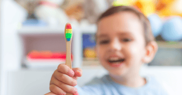 A small boy, faded in the background, holds up a rainbow toothbrush.