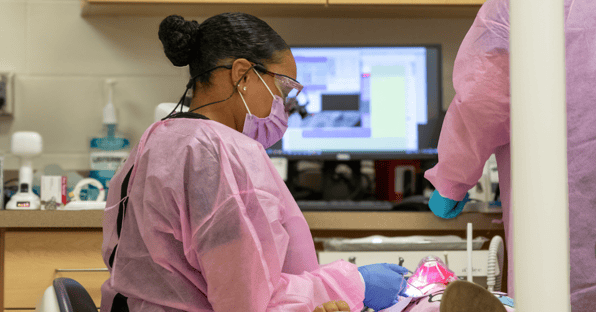 A Black woman wearing magnifying loupes, a purple mask and a pink protective gown provides dental care to a child who is out of view. 