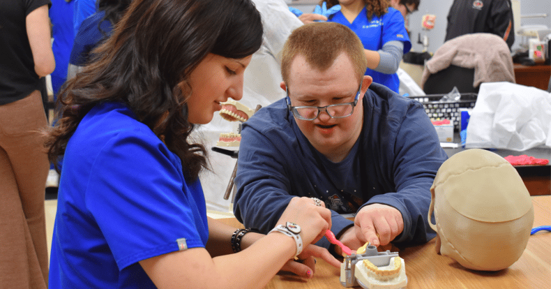 A young woman in blue scrubs works on a mouth model with a young man who has an intellectual and developmental disability.