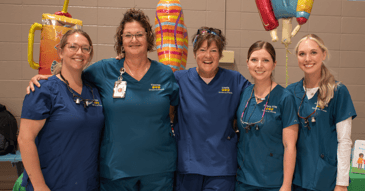 A Black woman wearing magnifying loupes, a purple mask and a pink protective gown provides dental care to a child who is out of view. 