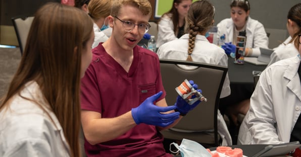 A male dental student wearing maroon scrubs holds up a mouth model as he talks to high school students.