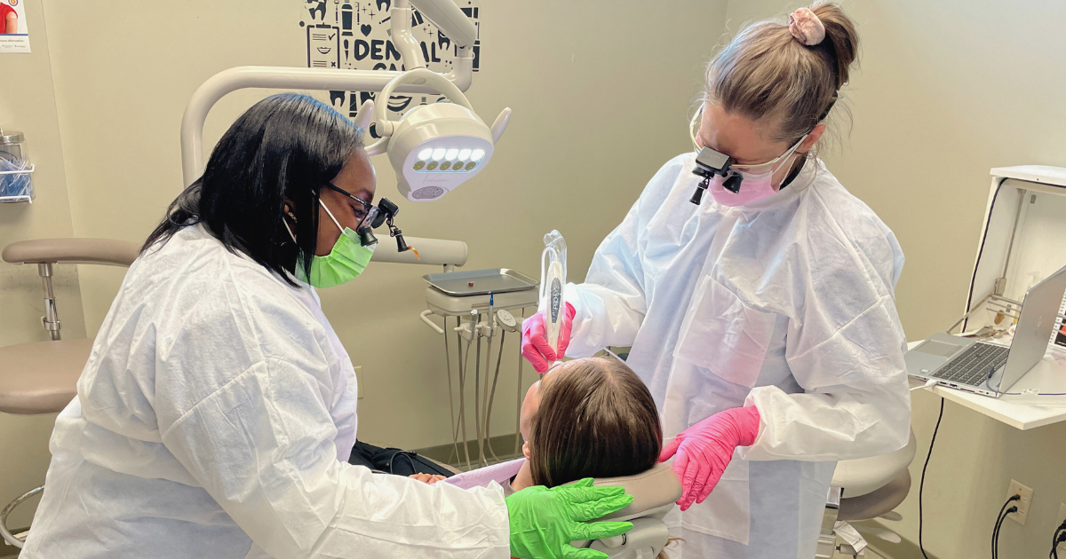 A female dentist and hygienist work with a patient seated in a dental chair facing away from the camera. One of the oral health professionals is Black, and the other is white. They both wear colorful nitrile gloves and matching masks.