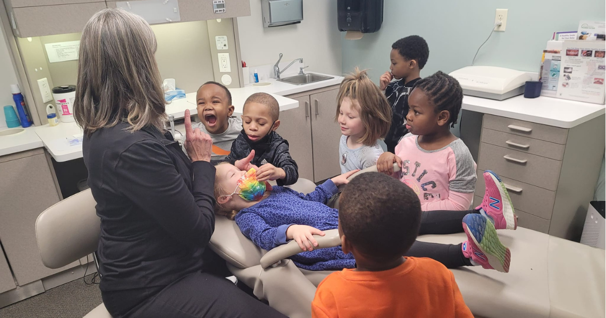 A hygienist with shoulder-length gray-brown hair faces away from the camera, one finger up in the air as she talks to a group of children under the age of 10. One child with a tie-dye mask reclines in a dental chair. The rest of the children gather around, watching intently. 