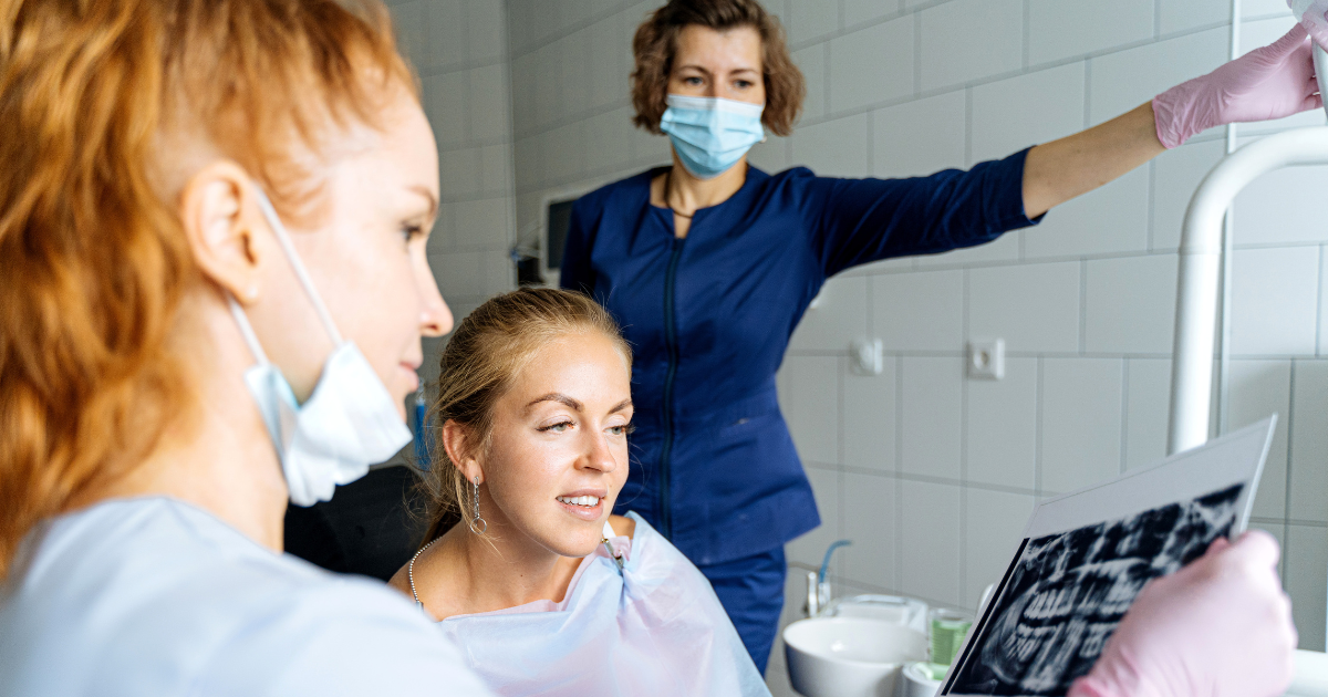 Three white-presenting women review a set of dental X-rays. One is a dentist, one is a hygienist, and one is a patient in the dental exam chair.