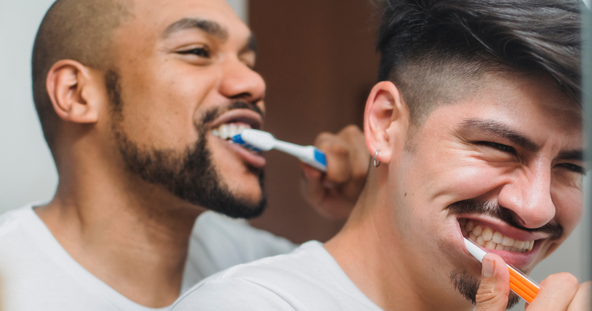Close-up photo of a Black man and a Hispanic man brushing their teeth together. 