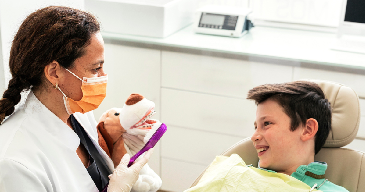 A Black female oral health professional demonstrates brushing techniques using an oversized toothbrush and a stuffed animal for a smiling boy in a dental chair.