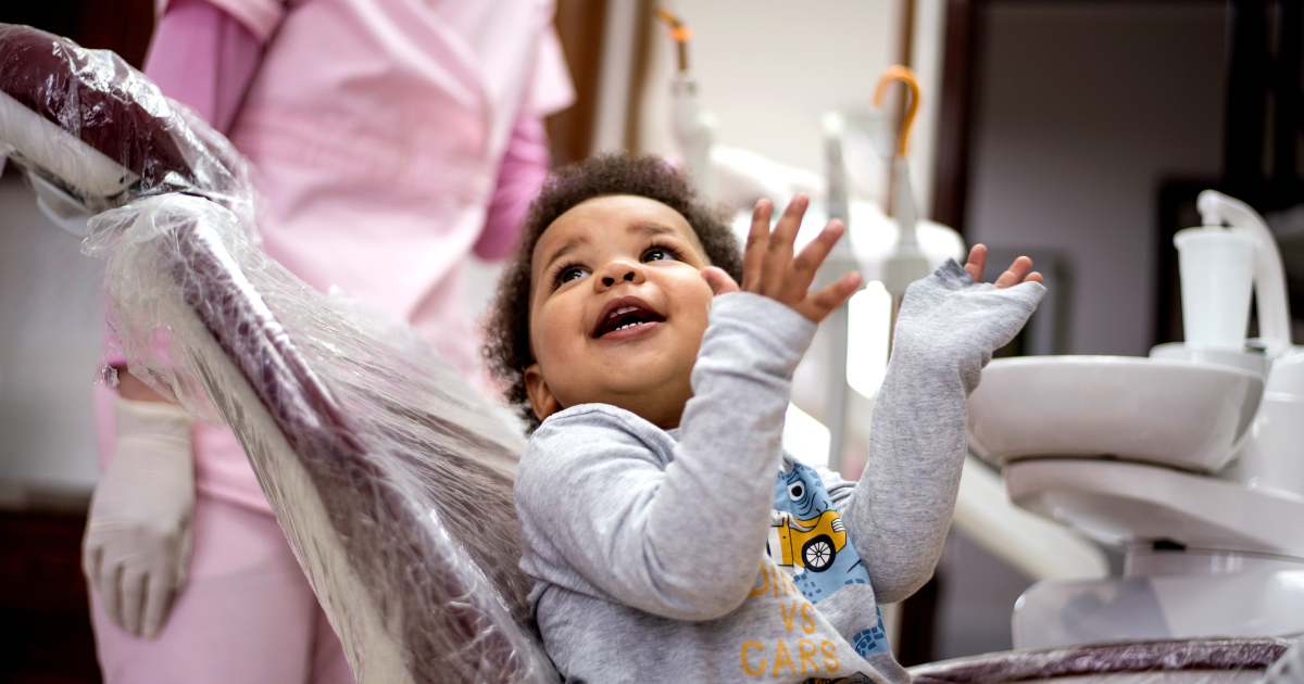 An African American child wearing an oversized gray longsleeved shirt smiles up at someone from the dental chair. Someone wearing pink scrubs stands in the background.